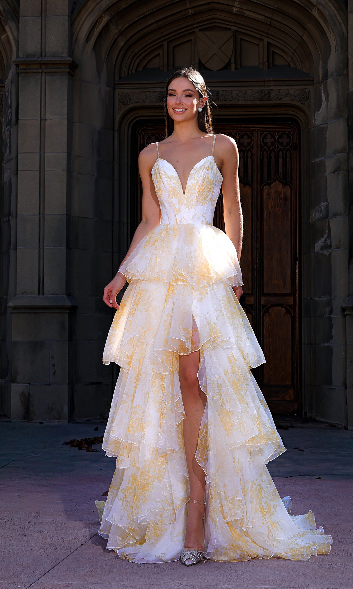 A woman stands outdoors before a stone building with an arched wooden door, wearing the Terani 261P6673 long formal dress in Yellow Multi—a printed chiffon design with thin straps and a tiered high-low silhouette.
